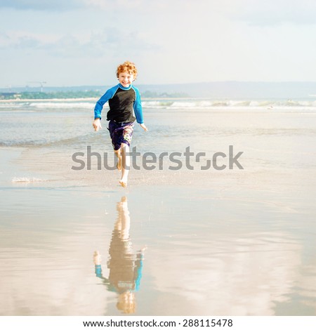 Happy boy in swimming shorts and rushwest runs along Bali beach near sunset with reflection in the water
