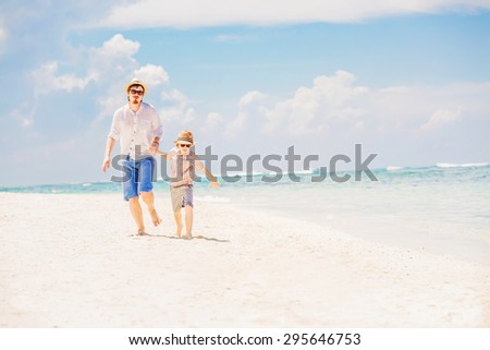 Father and son in summer nats and sunglasses running barefoot on the sea surf line having great holidays time on beautiful sunny day. Bali, Pandawa beach