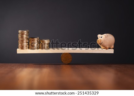 Seesaw with piggy bank on one side and stack of money on the other side.