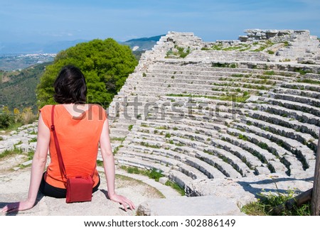 Tourist girl with an orange t-shirt sitting on one of the steps of the Segesta theater, West Sicily, and looking at the view