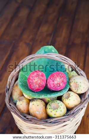 Red cut in half prickly pear on a cactus leave and a wicker basket full of just picked prickly pears on a wooden table