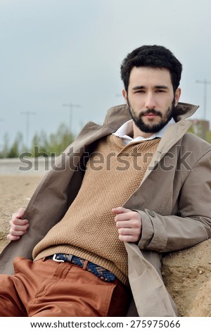 Handsome young man sitting in a coat on the sand in windy weather