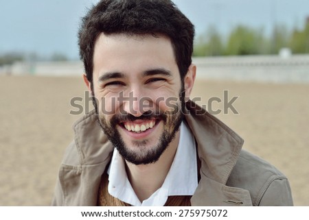 Handsome young man stands in a coat on the sand in windy weather and smiles