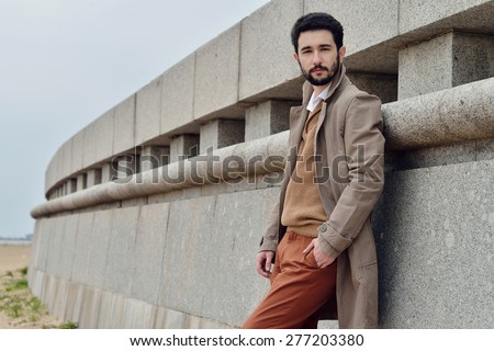 Handsome young man stands in a coat on the sand in windy weather