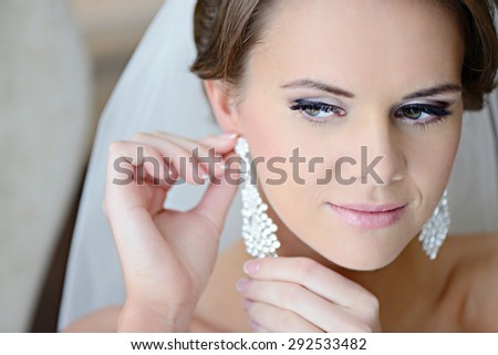 Beautiful bride in white wedding dress puts on earring. Beauty model girl is wearing jewelry. Female portrait in bridal gown for marriage. Woman with curly hair and lace veil. Cute lady indoors