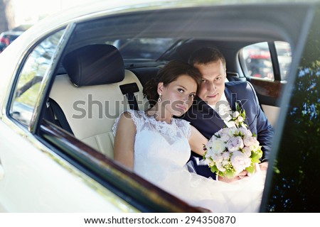 Wedding couple is hugging in a car. Beauty bride with groom. Beautiful model girl in white dress. Man in suit. Female and male portrait. Woman with lace veil. Cute lady and guy outdoors