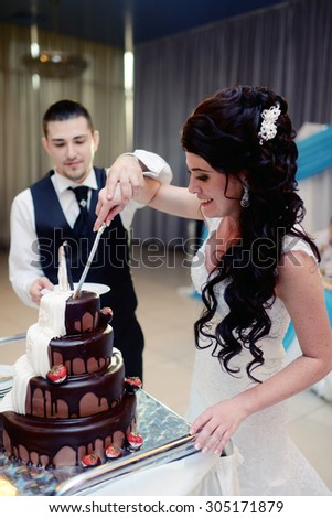 Beauty bride and handsome groom are cutting a wedding cake. Couple in the restaurant with colorful pie. Beautiful model girl in white dress. Man in suit. Female and male portrait. Cute lady and guy