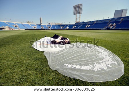 Wedding couple on the football stadium is hugging each other. Beautiful girl in white dress. Man in suit. Beauty bride with groom. Female and male portrait. Woman with lace veil. Lady and guy outdoors
