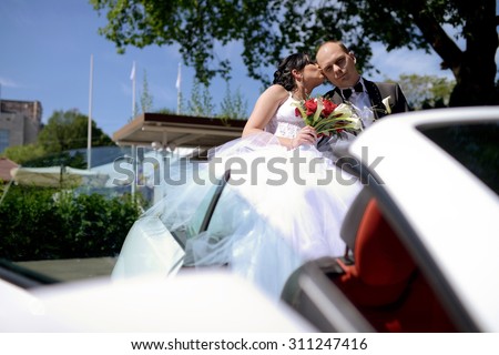 Wedding couple is hugging in a car. Beauty bride with groom. Beautiful model girl in white dress. Man in suit. Female and male portrait. Woman with lace veil. Cute lady and guy outdoors