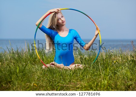 outdoor portrait of young beautiful blonde woman gymnast training with hula-hoop on green grass
