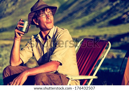 Colorized vintage outdoor portrait of tourist in camp sitting on the chair near the river and drinking tea