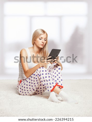 Young blonde woman in pyjamas on white whole-floor carpet reading e-book  near window