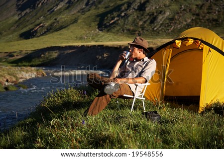 man is sitting in the chair at the camp in mountains near the stream