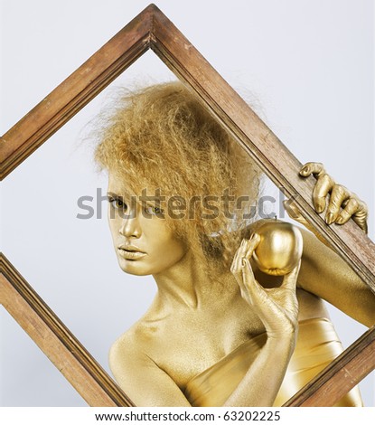 portrait of girl with golden bodyart posing with golden apple in her hands in picture frame on gray