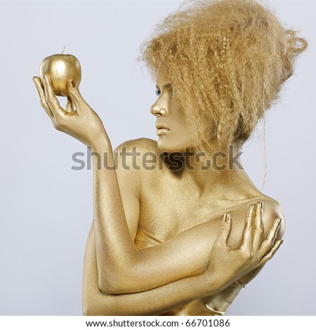 portrait of girl with golden bodyart posing with golden apple in her hands on gray