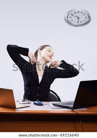 office portrait of beautiful tired business woman stretching at her workplace with two laptops on gray
