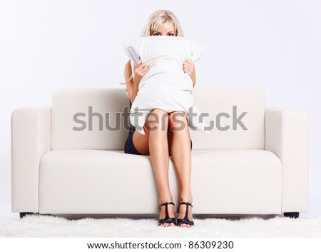 portrait of beautiful young blond woman sitting on couch with remote control watching scary movie and hiding behind the pillow