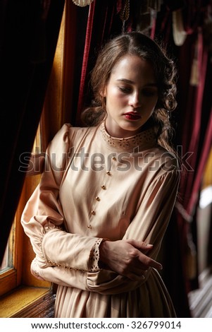 Young woman in beige vintage dress of early 20th century standing near window in corridor of retro railway train
