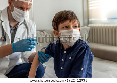 The smiling boy is looking away while his doctor holding a syringe next to his arm. Pediatrician makes vaccination to cute Caucasian boy. Boy in medical face mask getting flu shot by doctor at home. 