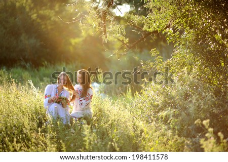 two girls in Ukrainian national dress sitting on the grass. Girl braids braid his girlfriend