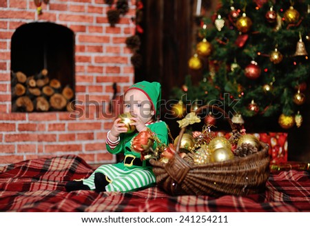 child dressed as an elf sitting on a plaid background on the Christmas tree and a brick fireplace. A child dressed as an elf gnawing teeth Christmas toy.