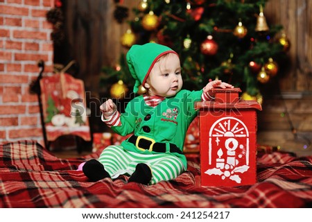child dressed as an elf sitting on a plaid background on the Christmas tree and a brick fireplace. A child dressed as an elf stretches his hand to the lamp