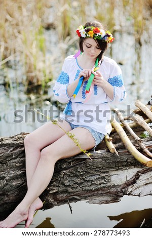 Ukrainian girl in a dress and a wreath of flowers on her head sitting on a log by the river
