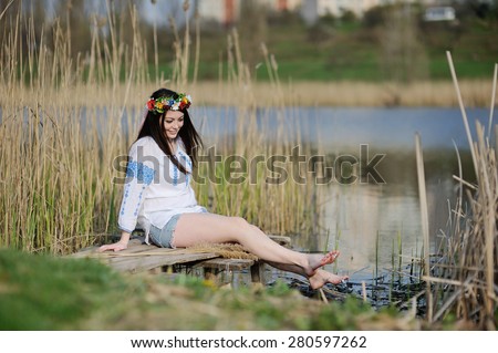 girl wets feet in the river. girl sitting on the bridge in the Ukrainian shirt and a wreath of flowers and ribbons on the head