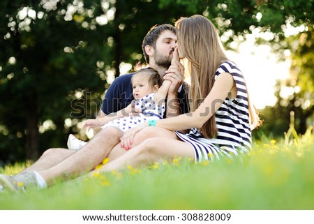 Young family relaxing in the park on the grass. Mom and dad kiss his hand a small daughter