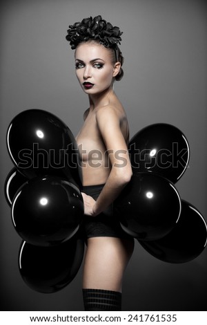 Beautiful fashion model with perfect skin posing topless covered by black balloons, with great makeup, mascara, lipstick, eyelashes, eyebrows hairstyle and black flower in her long hair