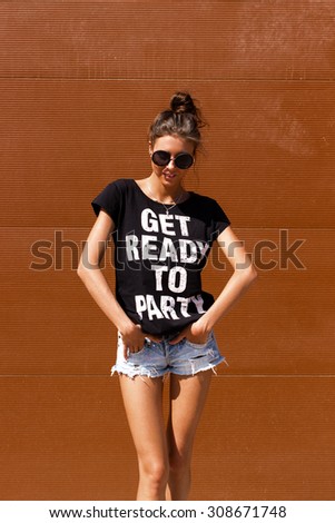 Young European teenage brunette curly model posing near brown ribbed wall, grimacing, jumping, wearing blue jeans mini shorts, sneakers, round sunglasses and black t-shirt, ready to party
