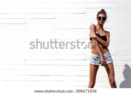 Young European teenage brunette curly model posing near white wooden wall, grimacing, jumping, wearing blue jeans mini shorts, sneakers, round sunglasses and black swimwear top