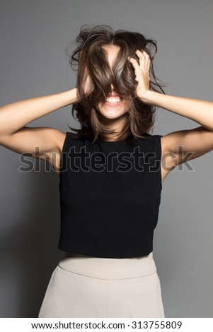 Awesome Caucasian attractive joyful happy sexy female model is shaking head with brunette hair in studio wearing black sleeveless shirt, isolated on gray background
