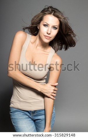 Awesome Caucasian attractive joyful happy sexy female model is shaking head with brunette hair, grimacing in studio, wearing beige sleeveless shirt, isolated on gray background