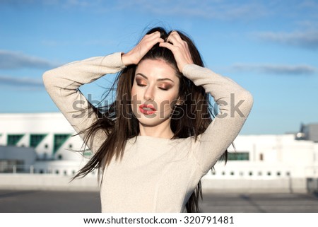 Attractive young brunette woman with pretty makeup shaking head, touching hair by her hands, standing outdoors on a bright sunny day in front of buildings under the blue sky