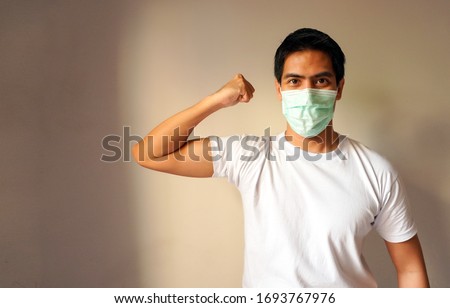 Portrait of a handsome Asian man wearing medical protective mask standing with a raised arm shows strong biceps. Isolated background. Concept of staying strong and healthy against coronavirus covid-19