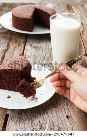 Woman\'s hand picking chocolate cake on white plate. Glass of milk and chocolate cake are background. Over wooden table.