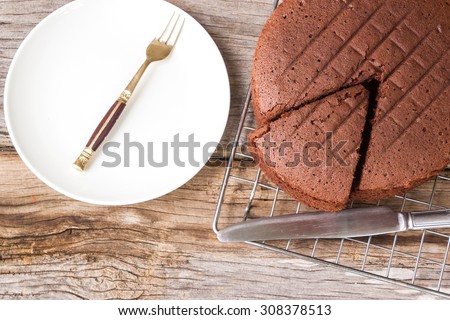 Chocolate cake on baking rack with knife. And fork on white plate. Over wooden table. Top view.