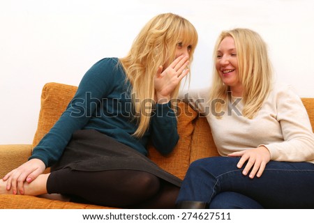 2 blond girl friends gossiping,  whispering and telling a shocking secret and smiling sitting on a sofa at home