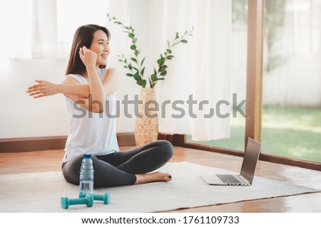 Smiling Asian woman doing yoga shoulder stretching online class from laptop at home in living room. Self isolation and workout at home during COVID-19.