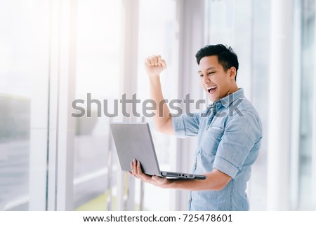 Happy excited Asian man holding laptop and raising his arm up to celebrate success or achievement. 商業照片 © 