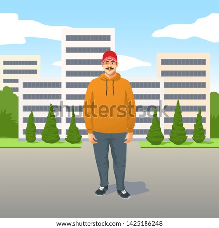 Relaxed trendy young Latino man with mustache in casual clothes and a baseball cap standing in the road in front of high-rise urban apartment blocks