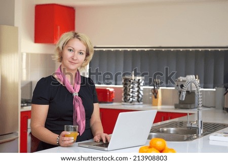 Blonde woman in black dress is working on a laptop in a red kitchen.