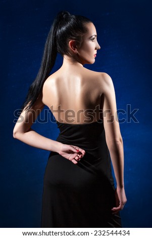 Brunette girl with long ponytail in a black dress on a dark blue background posing back