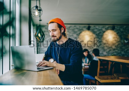Caucasian male resting in coffee shop listening favorite music in headphones and chatting in social networks, skilled freelancer enjoying remote job typing publication for web blog on laptop