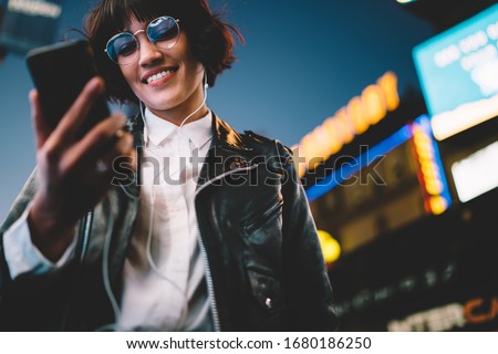 Below view of pretty woman in cool eyeglasses and trendy wear smiling on metropolitan street with night lights enjoying songs from playlist in earphones, girl reading sms with good news on smartphone