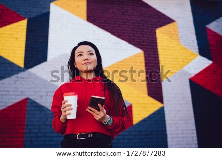 Pensive Asian youngster with takeaway cup and mobile phone thinking at urban setting with street art, trendy dressed generation Z holding cellular gadget and coffee to go spending leisure in city Stok fotoğraf © 