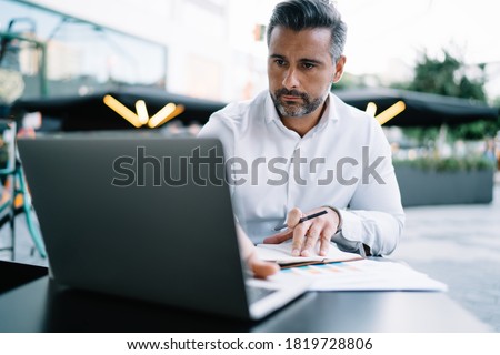 Mature Caucasian corporate director analyzing web information during organisation planning in street cafeteria, middle aged businessman reading news via netbook technology connected to 4g wireless