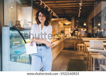 Half length portrait of prosperous manager of local bakery smiling at camera during work day in franchise takeaway cafe, happy woman with paper statistics enjoying time for improve own coffeehouse