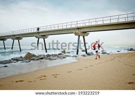 Young athlete man in white t-shirt and sports a red windbreaker runs under the pier along the sea